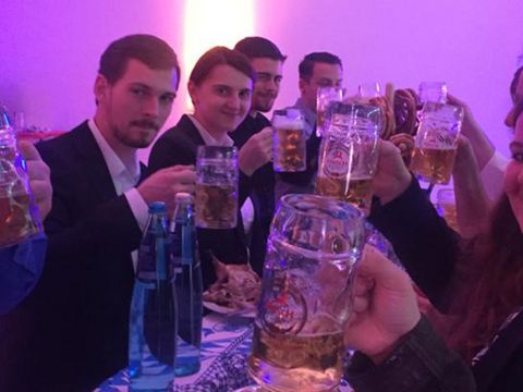 Gruppe von Mitarbeiter sitzen auf Bierbänken und stoßen mit Bier an alle lächeln in die Kamera