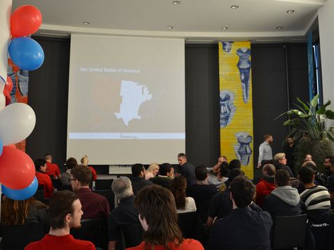 Viele Personen sitzend vor einer kleinen Bühne mit einer Leinwand